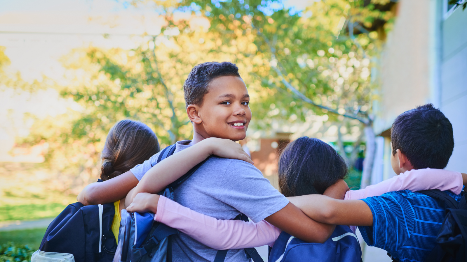 Four smiling students standing outdoors with their arms around each other’s shoulders, wearing backpacks.